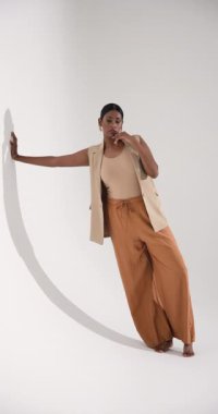 Dikey video: Moda stüdyosunda zarif bir şekilde beyaz duvara poz veren şık giyimli bir model. Modellik, zarafet, tarz, fotoğraf çekimi, giysi