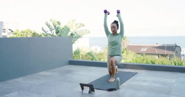 Woman following prompts stepping onto mat on terrace and doing dumbbell lunge press for fitness. Fitness, outdoor, minimalist, well-being, strength, lifestyle, training