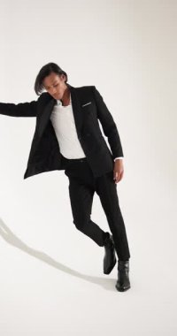 Dikey video: Siyah takım elbiseli şık adam beyaz stüdyo duvarına karşı güvenle poz veriyor. Moda, güven, erkek giyim, zarafet, entelektüellik, modellik.