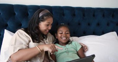 Anne tableti alıyor ve ekranı paylaşıyor, kızına yatakta görüntülerde rehberlik ediyor. Aile, bağlantı, eğitim, teknoloji, konforlu, bağlanma, boş zaman