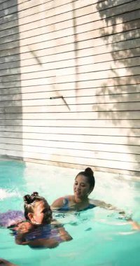 Dikey video: Anne ve kızı havuzda neşeyle yüzüyor, güneşli bir öğleden sonranın tadını çıkarıyorlar. Aile, kaynaşma, yaz, eğlence, eğlence, açık hava.