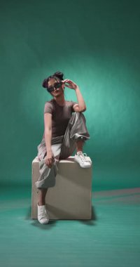 Dikey video: Gündelik giyinen şık bir kadın küpün üzerinde oturuyor, güneş gözlüğünü güvenle tutuyor. Moda, güven, tarz, moda