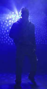 Dikey video: Deri ceketli özgüvenli adam dramatik mavi stüdyo ışıkları altında poz veriyor. Moda, güven, kentsel, tarz, modern, erkek