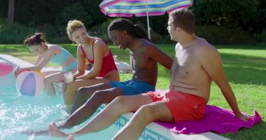 Havuz kenarına gelen farklı arkadaşlar havuz kenarında oturup sosyalleşmek için ayaklarını plaj topuna daldırıyorlar. Yaşam tarzı, rahatlama, rahatlama, açık hava, canlı, yoldaşlık, gündelik