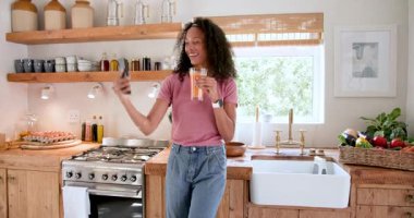 Sıcak mutfakta taze meyve suyu içen, gülümseyen ve akıllı telefon tutan bir kadın. İçecek, yaşam tarzı, teknoloji, rahatlama, su, beslenme