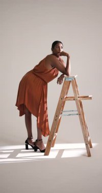 Dikey video: Akan elbiseli zarif bir kadın stüdyoda tahta merdivenin yanından güvenle geçiyor. Moda, zarafet, tarz, güven, model, cazibe