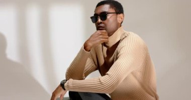 Başlangıçta Afrika kökenli Amerikalı bir adam güneş gözlüğünü ayarlıyor. Stüdyo sandalyesinde modellik için kol saatini kontrol ediyor. Portre, tefekkür, moda, minimalizm, sofistike, zarafet, profesyonel