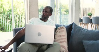 Afro-Amerikalı adam oturma odasında kanepede oturuyor, akıllı telefon görüşmeleri yapıyor ve dizüstü bilgisayar yazıyor. Modern, çalışma alanı, iletişim, üretkenlik, teknoloji, stil, minimalist