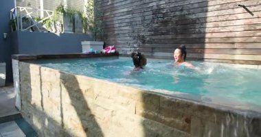 Arka bahçedeki platformda gülen Asyalı anne ve mayo giymiş Afrikalı Amerikalı kız havuza atlıyorlar. Aileler, eğlence, neşe, eğlence, eğlence, açık hava, kaynaşma.