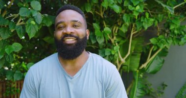 Afro-Amerikalı adam bahçede kollarını kavuşturarak duruyor, yemyeşil yaprakların arasında gülümsüyor. Kendinden emin, rahat, yeşillik, açık hava, doğa, mutluluk, yaşam tarzı