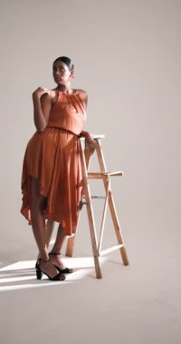 Dikey video: Turuncu elbiseli zarif kadın stüdyodaki tahta merdivende güven içinde poz veriyor. Moda, portre, zarafet, tarz, özgüven, kıyafet.