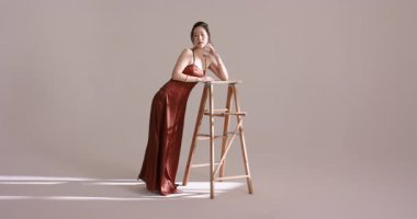 Fotoğrafçı, Asyalı kadın modelin bordo elbisesini göstermek için stüdyoda merdivene yaslandığını söylüyor. Zarafet, sofistike, cazibe, portre, minimalist, moda, güzellik.