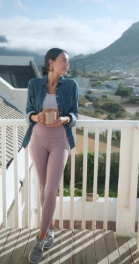 Dikey video: Balkonda sabah kahvesinin keyfini çıkarmak, aktif giyimli kadın huzurlu manzaranın tadını çıkarıyor. Gün doğumu, rahatlama, farkındalık, sükunet, kendine bakım, tefekkür