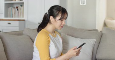 Asyalı kadın süprizi fark ettikten ve evdeki açık gri koltukta dinlendikten sonra akıllı telefonu kaydırıyor. Rahat, rahat, güncel, çağdaş, iç mekan, yaşam tarzı, konfor