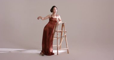 Çinli kadın model stüdyoya giriyor, merdivene yaslanıyor, geri dönüyor, yanaklarını ışığa sürtüyor. Zarif, moda, cazibe, minimalizm, dengeli, güzellik, sofistike