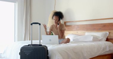 Yatakta dizüstü bilgisayar ve telefon kullanan bir kadın, yanında bavul, gülümseyerek, evde. Seyahat, teknoloji, iletişim, yaşam tarzı, rahatlık, konaklama