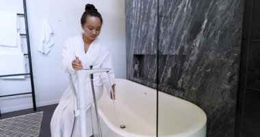 Bornozlu yetişkin bir kadın küvet musluğunu değiştiriyor, sıcaklığı test ediyor evde banyo hazırlıyor, fotokopi çekiyor. Lüks, rahatlama, kaplıca, modern, refah, konfor, minimalizm