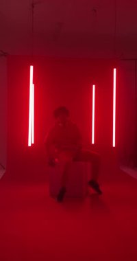 Dikey video: Kırmızı elbiseli bir adam stüdyoda canlı kırmızı ışıklar altında küpe yaslanıyor. Moda, portre, tarz, yaratıcılık, yenilikçi, canlı