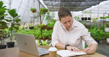 Serada dizüstü bilgisayarı ve kahvesi olan bir kadın deftere yazı yazıyor. Bahçecilik, bahçıvanlık, teknoloji, not alma, kafein, çalışma alanı