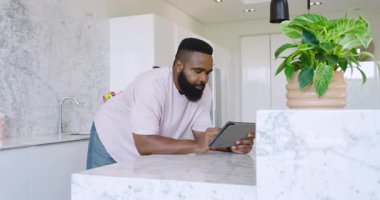 Modern mutfakta mermer tezgahın üzerine eğilip tablet cihazını kontrol eden Afro-Amerikalı bir adam. Modern, mutfak, teknoloji, bağlılık, çağdaş, minimalizm, odaklanma