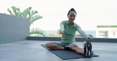 Orta yaşlı Asyalı kadın, yoga minderi ve su şişesiyle çatıda hareket edebilmek için bacak esneme hareketleri yapıyor. Fitness, sağlık, hareketlilik, açık hava, yaşam tarzı, rahatlama, gençleşme