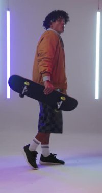 Dikey video: Gündelik kıyafetli genç adam modern stüdyoda kaykay tutuyor. Kaykay, moda, yaşam tarzı, şehir, güven, gençlik