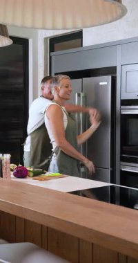 Dikey video: Olgun çift mutfakta birlikte yemek hazırlarken neşeyle dans ediyor. Yemek pişirme, ilişki, mutluluk, birliktelik, yaşam tarzı, yerli