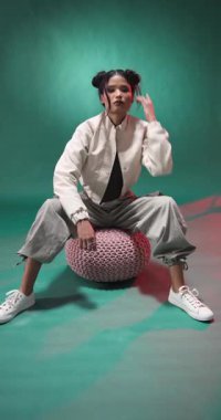 Dikey video: Şık giyinmiş genç bir kadın puf üzerinde oturuyor, grunge modasını benimsiyor. alternatif, keskin, trend, kentsel, giyim, yaşam tarzı