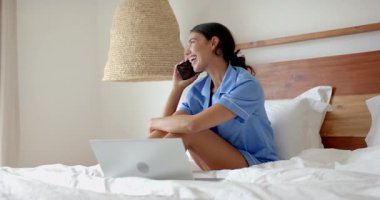 Mavi pijamalı bir kadın dizüstü bilgisayar kullanarak yatakta oturuyor ve evde telefonla konuşuyor. Uzaktan çalışma, teknoloji, iletişim, yaşam tarzı, çoklu görev, ev ofisi