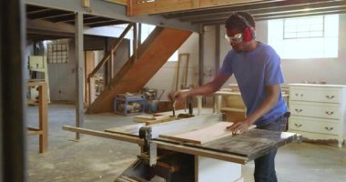 Orta siyah erkek marangoz atölyeye girip masa testeresiyle tahta kesiyor. Sanatçılık, ahşap işçiliği, işbirliği, sanayi, kesinlik, atölye, aletler