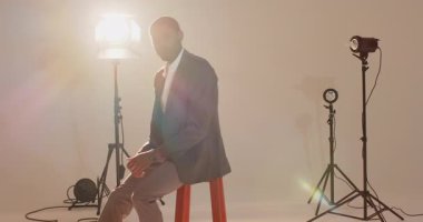 Afro-Amerikalı adam taburede poz veriyor, ceketin yakasını çekiyor ve stüdyo çekimi için nöbet tutuyor. Profesyonel, moda, portre, stüdyo, erkek, tarz, zarafet