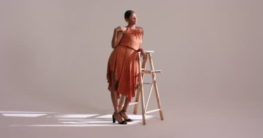 Hintli kadın model, yanmış turuncu elbiseyi gösteren güneş ışığı altındaki stüdyoda merdiven üzerinde poz veriyor. Moda, zarafet, minimalist, ticari, yaratıcı, stüdyo, güzellik