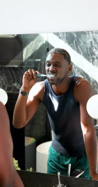 Dikey video: adam banyoda dişlerini fırçalıyor, aynaya bakıyor, sabah rutini. Hijyen, bakım, cilt bakımı, yansıma, kişisel bakım