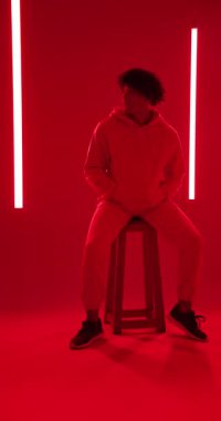 Dikey video: kapüşonlu genç adam canlı kırmızı neon ışıkların altında taburede oturuyor. Şehir, gece hayatı, modern, moda, karamsar, gençlik