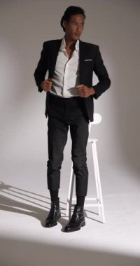 Dikey video: kendine güvenen şık takım elbiseli adam taburede oturuyor, stüdyoda ceketini ayarlıyor. Moda, özgüven, zarafet, resmi giyim, tarz, bakım