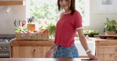 Kırmızı tişörtlü gülümseyen kadın güneşli mutfağın tadını çıkarıyor, pencereden dışarı bakıyor, evde. Güneş ışığı, mutluluk, sabah, yaşam tarzı, sıradan, huzurlu