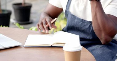 Dizüstü bilgisayarda yazan, akıllı telefon ve defterle çalışan Afro-Amerikan bir adam, seranın yakınlarında. Teknoloji, dijital, çoklu görev, çalışma alanı, odaklanma, üretkenlik