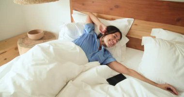 Mavi pijamalı bir kadın yatakta uyanıyor, gülümsüyor ve evinde mutlu bir şekilde esniyor. Sabah, rutin, rahatlama, mutluluk, sağlık, yatak odası.