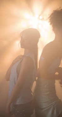 Dikey video: İki kadın dramatik ışıklandırmada güven içinde poz veriyor, cesur moda tarzını ifade ediyorlar. cazibe, güven, tutum