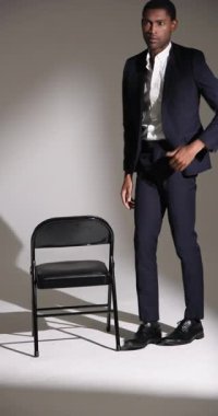 Dikey video: Şık takım elbiseli kendine güvenen adam sandalyede oturuyor, stüdyoda düşünüyor. Moda, iş, profesyonel, modern, güven, tarz