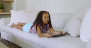 Propping elbows, school-age girl tapping and rotating tablet on living room sofa, discovering game. Children, digital, modern, entertainment, relaxation, learning, technology