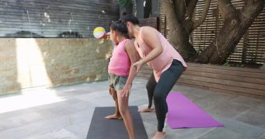 Farklı anne ve kız teras paspasları üzerinde Savaşçı II pratiği, yol gösterici duruş ve gülümseme paylaşımı. Yoga, sağlık, aile, açık hava, uyum, farkındalık, fitness