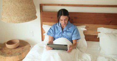 Yatakta gülümseyen kadın tablet kullanıyor, mavi pijama giyiyor, evde sabahın tadını çıkarıyor. Teknoloji, rahatlama, rahatlama, yaşam tarzı, dijital cihaz, sabah rutini
