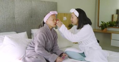 Asyalı anne-kız ürünlerin ambalajlarını açıyor, krem sürüyor ve kırmızı göz altı jel bantları yapıştırıyor. Cilt bakımı, aile, şefkat, sağlık, rahatlık, samimiyet, sükunet.