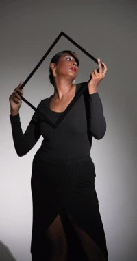 Dikey video: Siyah elbiseli kendine güvenen kadın moda stüdyosunda boş bir çerçeveyle poz veriyor. yaratıcılık, zarafet, tarz, portre, tasarım, lüks