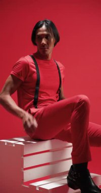 Dikey video: Kırmızı elbiseli manken stüdyodaki beyaz kutuda güvenle oturuyor. Modellik, moda, şık, tarz, zarafet