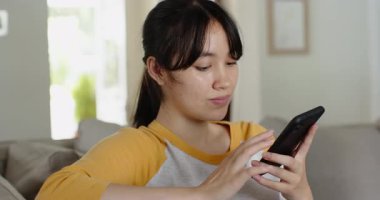Asyalı kadın akıllı telefondan içeriğini kaydırıyor, gülümsüyor, sesli mesaj gönderiyor ve evdeki kanepede dinliyor. Gündelik, yaşam tarzı, modern, iletişim, rahatlık, konfor, doğal