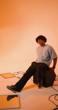 Dikey video: Stüdyoda kablolar ve mikrofonla çevrili bir amplifikatörün üzerinde oturan genç adam. Müzik, müzisyen, kayıt, ses, ses, teknoloji