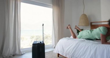 Kadın yatakta dinleniyor, akıllı telefon kullanıyor, bavul yakınlarda tatilin tadını çıkarıyor, evde. Seyahat, rahatlama, rahatlama, kaçamak, dalgınlık, otel