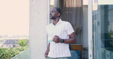 Terasta dikilen, kahve fincanından yudumlayan ve yeşillik ve dekora bakan Afro-Amerikalı bir adam. Balkon, yeşillik, kahve, tefekkür, modern, açık hava, rahatlama.